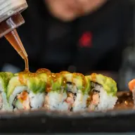 En sushikock ringlar sås över maki med avokado medan inlagd ingefära ligger bredvid på plåten.