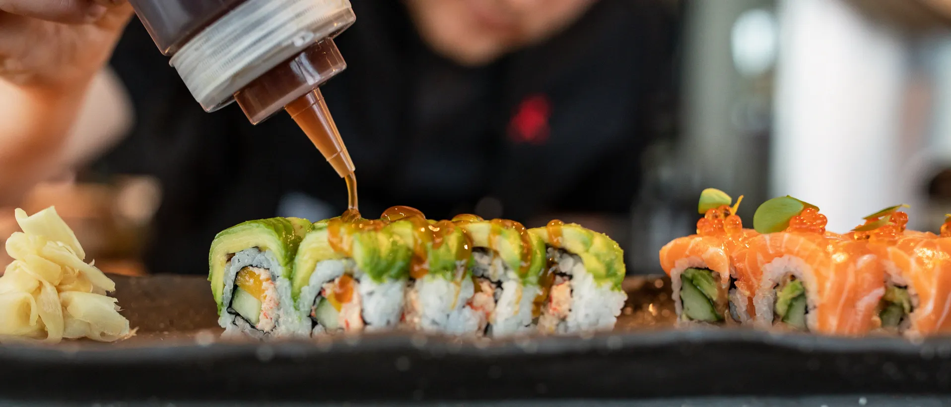 En sushikock ringlar sås över maki med avokado medan inlagd ingefära ligger bredvid på plåten.